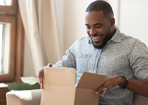 Happy african american man sit on couch feel excited opening cardboard box delivery parcel, smiling biracial millennial male shopping online unpack unbox online order, overjoyed with service or goods